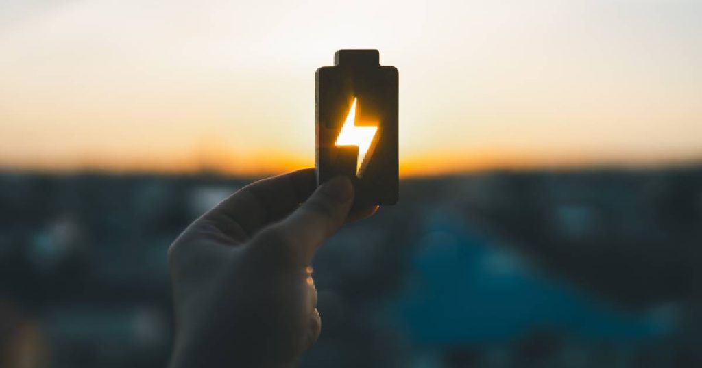 Een hand met een voorstelling van een batterij waaruit een bliksemschicht gesneden is. Op de achtergrond de zonsondergang. Het zonlicht schijnt door de uitsnijding in de batterij. 
