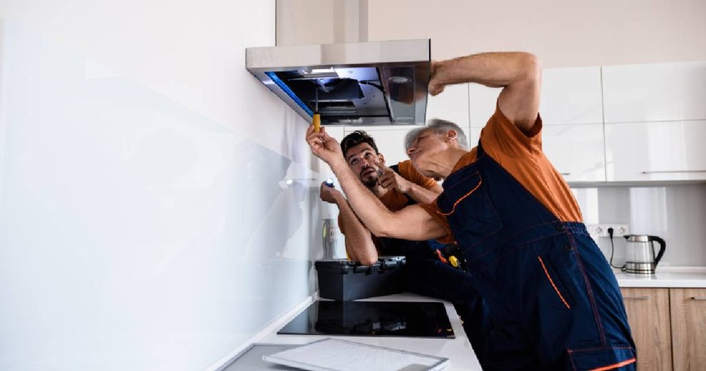 Twee mannen in blauwe overalls en oranje T-shirts zijn een dampkap aan het vervangen tijdens het zelf renoveren van de keuken.