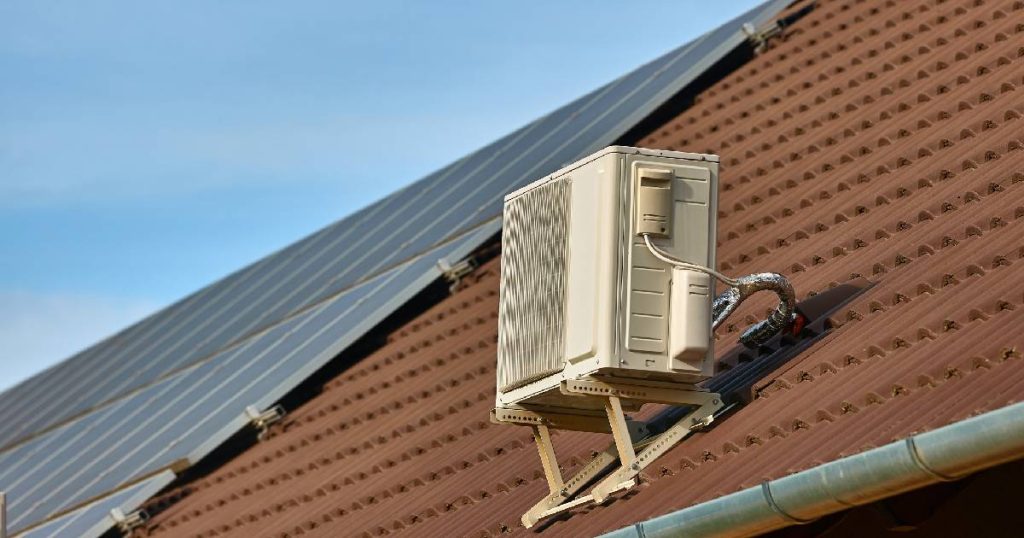 Een hellend, roodbruin dak met dakpanelen, donkerblauwe zonnepanelen en de geelwitte buitenunit van een vast airconditioning systeem.