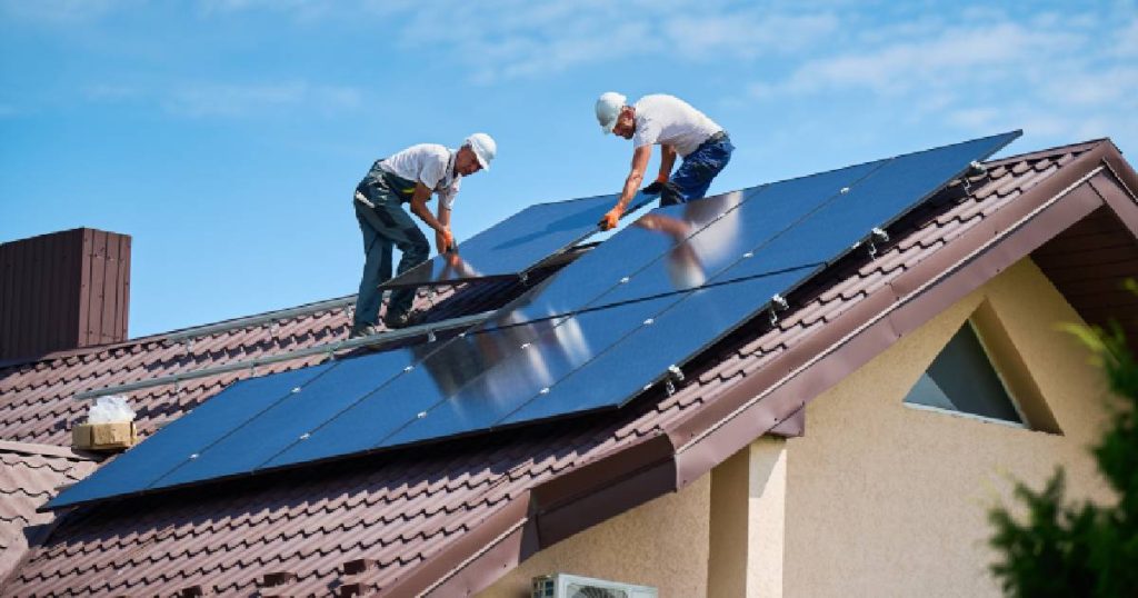 Twee installateurs leggen nieuwe zonnepanelen op het pannendak van een huis. Ze dragen elk een jeansbroek, wit T-shirt en witte veiligheidshelm.