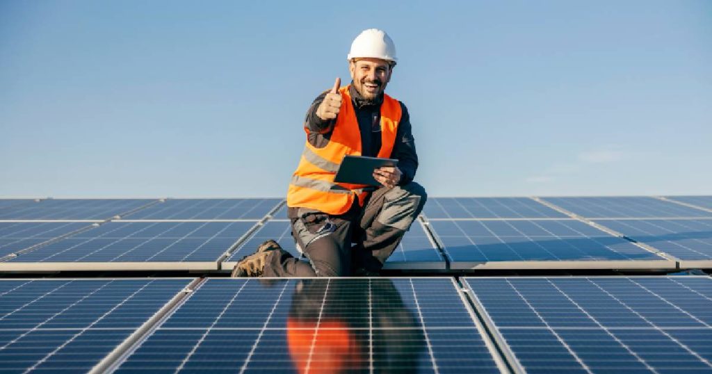 Een zonnepaneelspecialist na een positieve keuring van een aantal zonnepanelen. Hij draagt een oranje fluohesje, witte helm, zwartgrijze werkbroek en zit geknield tussen enkele zonnepanelen. Hij steekt zijn duim op en glimlacht.